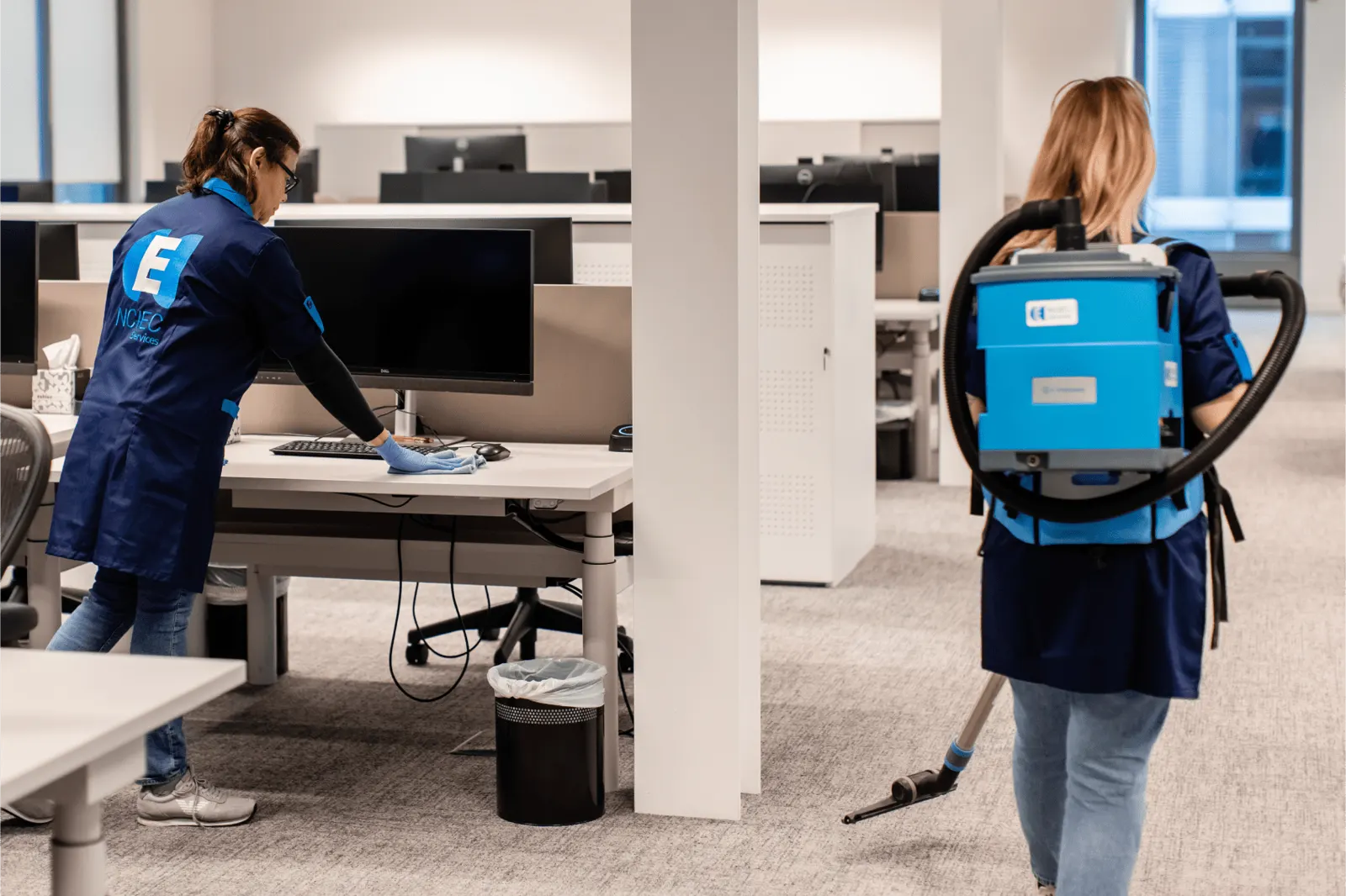 Deux agentes NCIEC nettoient un plateau de bureaux ouvert, l'une avec un aspirateur dorsal